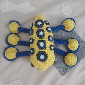 May include: A crocheted yellow and blue octopus with white and blue polka dots. The octopus has six tentacles and is lying on a gray surface.