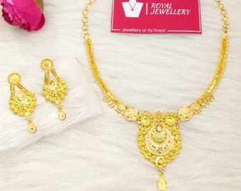 May include: A gold necklace and a pair of matching gold earrings. The necklace features a central pendant with intricate filigree and a small teardrop charm. A pink rectangular card with the white text Royal Jewellery is positioned above.