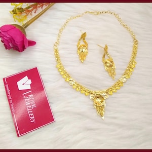 May include: A gold-toned necklace and matching pair of drop earrings featuring intricate textured metalwork. The necklace has a central floral-inspired pendant with dangling elements. A pink card with the text Royal Jewellery is included.