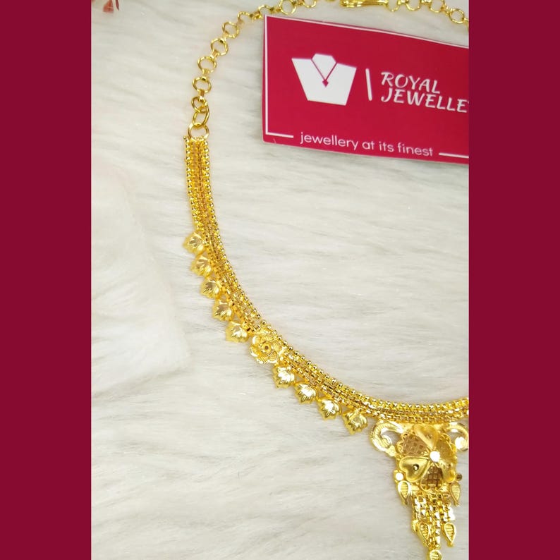 May include: A gold chain necklace featuring a series of small, dangling leaf-shaped charms along the length and a larger, intricate floral-inspired pendant at the center. A red tag with the white text ROYAL JEWELLE is positioned near the chain.