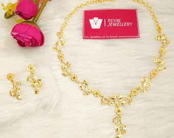 May include: A gold-toned necklace and matching drop earrings set featuring a delicate floral and leaf motif. The necklace includes a central pendant element. A small rectangular pink card with the text Royal Jewellery is positioned above.