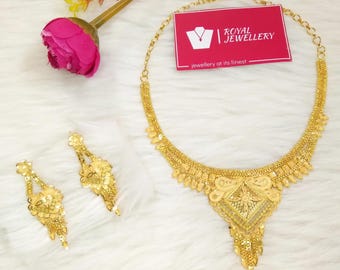 May include: A gold-toned necklace and matching pair of drop earrings featuring intricate filigree and textured metalwork. A pink artificial flower and a rectangular pink card with the white text Royal Jewellery are positioned near the jewelry set.