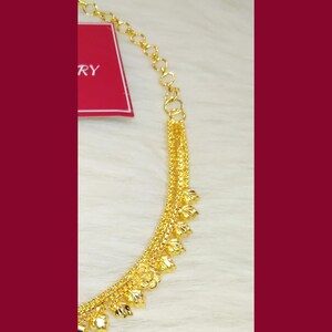 May include: A gold-toned necklace featuring a delicate chain with a series of small, textured leaf-shaped charms hanging along the lower edge. A red rectangular tag with the partial white text RY is attached to the upper portion of the chain.