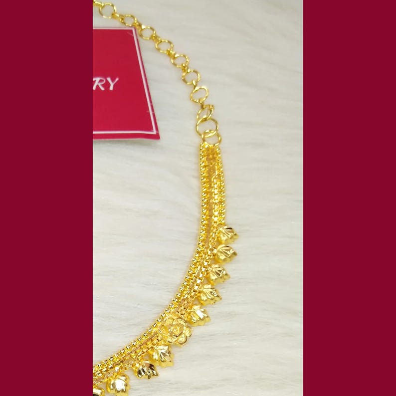 May include: A gold-toned necklace featuring a delicate chain with a series of small, textured leaf-shaped charms hanging along the lower edge. A red rectangular tag with the partial white text RY is attached to the upper portion of the chain.