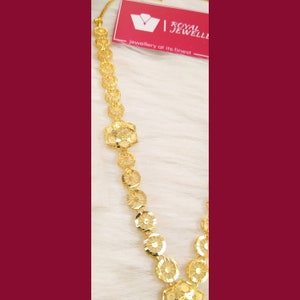 May include: A gold necklace with a delicate design featuring a series of interconnected circular and hexagonal shapes. The necklace is made of gold and has a shiny finish. The text "ROYAL JEWELLERY at its finest" is visible in the background.
