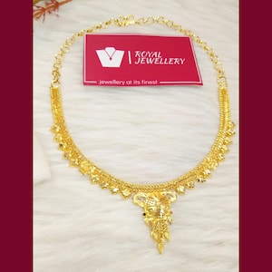 May include: A gold necklace featuring a delicate chain with small leaf-shaped charms and a central floral pendant. A rectangular red card is positioned behind the necklace, displaying the white text Royal Jewellery and jewellery at its finest.