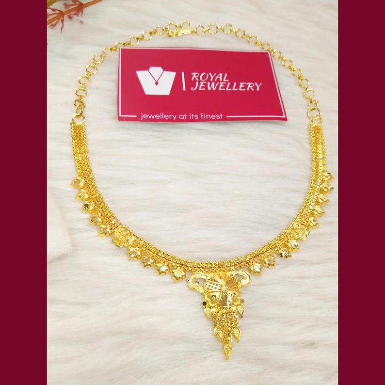 May include: A gold necklace featuring a delicate chain with small leaf-shaped charms and a central floral pendant. A rectangular red card is positioned behind the necklace, displaying the white text Royal Jewellery and jewellery at its finest.