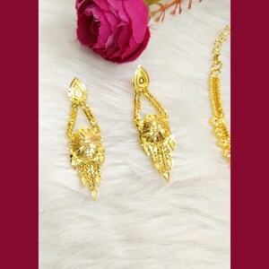May include: A pair of gold-toned drop earrings featuring a triangular top section connected to a circular floral motif with dangling chain fringe elements, accompanied by a partial view of a matching gold-toned chain necklace.