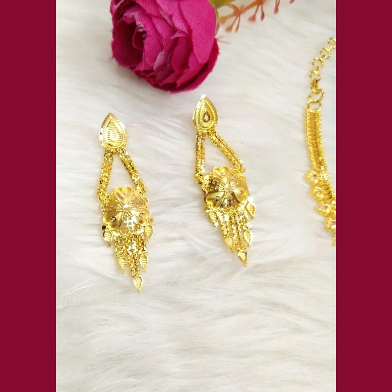May include: A pair of gold-toned drop earrings featuring a triangular top section connected to a circular floral motif with dangling chain fringe elements, accompanied by a partial view of a matching gold-toned chain necklace.