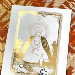 May include: A gold foil print of a Sikh Guru, with a white background and a gold ornate frame. The Guru is sitting with his hands clasped in front of him, and there is a halo around his head. The print has the symbol "ੴ" in the top right corner.