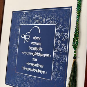 May include: A framed print with a blue background and silver lettering. The print features a traditional Sikh prayer in Gurmukhi script. The frame is surrounded by a decorative border of silver flowers. A green beaded mala hangs on the right side of the frame.