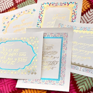 Op de afbeelding: Vier decoratieve papieren kaarten met sierlijke gouden en blauwe randen en ingewikkelde kalligrafie in goud en blauw. De kaarten liggen op een patroonachtige stoffen achtergrond.