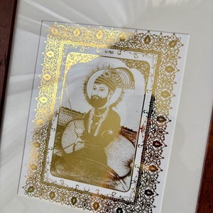 Peut inclure: Œuvre d'art religieuse encadrée avec une illustration dorée d'une figure avec un halo, portant un turban et tenant une épée. L'image est placée sur un fond blanc et encadrée de détails dorés ornés. Le cadre est de couleur marron foncé.