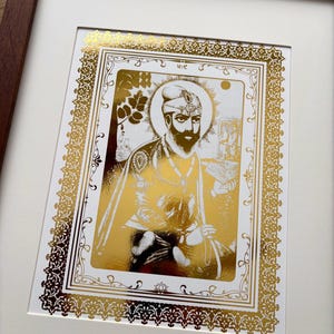 Op de afbeelding: Een ingelijste goudfolieprint met een portret van een bebaarde figuur met een tulband en traditionele kleding. Het kunstwerk bevat een gedetailleerde decoratieve rand, een halo-effect en een klein boek in de hand, alles tegen een witte achtergrond.