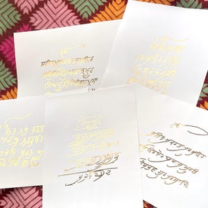 May include: Four white cards with gold calligraphy on them. The calligraphy is in a variety of styles and includes both English and non-English characters. The cards are arranged in a staggered pattern.