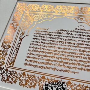 May include: A framed wall art piece featuring a white background with a metallic gold foil border and central text in Gurmukhi script. The intricate floral patterns frame the block of text, all encased within a light-coloured wooden frame.