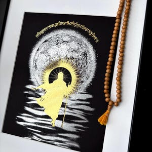 May include: Framed artwork with a gold figure and text on a black background, set against a white mount and black frame. A strand of wooden prayer beads with a yellow tassel hangs on the right. The artwork has a celestial theme.