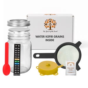 Puede incluir: Un tarro de cristal transparente con tapa metálica, una cuchara roja y una pegatina de termómetro. También hay una caja blanca con el texto "WATER KEFIR GRAINS INSIDE" y un colador negro. Una tapa amarilla y un pequeño paquete de granos de kéfir completan el conjunto.