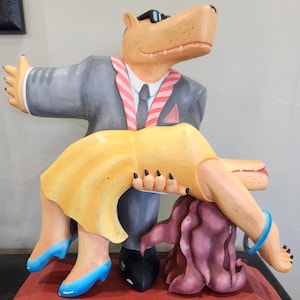 May include: A whimsical sculpture featuring a hippo in a suit and sunglasses holding a woman in a yellow dress. The sculpture is mounted on a rectangular base with the text "25/250 © 1987".