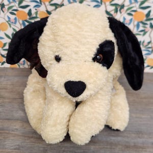 May include: A plush dog toy with cream-colored, textured fur. The stuffed animal features long, soft black ears, a black patch over one eye, and a small black nose. The toy is designed in a seated position with front paws resting forward.