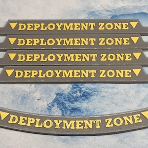 May include: Five gray plastic strips with the text "DEPLOYMENT ZONE" printed in yellow. The strips are arranged in a stack. Below the stack is a curved gray plastic strip with the same text printed in yellow.