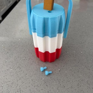 May include: A cylindrical container shaped like a tri-colored popsicle with a light blue top, white middle, and red bottom section. It features a light blue handle, a brown stick-like protrusion at the top, and two small light blue cylindrical pegs.