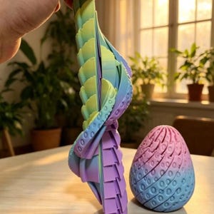 May include: A 3D printed articulated dragon figure with a gradient of green, blue, and purple, featuring a segmented body and textured scales. Beside it sits a matching 3D printed egg with a textured, patterned surface in pink, blue, and purple.