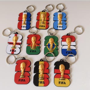 May include: A collection of eleven FIFA 2026 keychains, each featuring a gold-colored trophy figure centered on a rectangular base shaped like the number 26. Each keychain displays a different national flag design and the text FIFA in black.