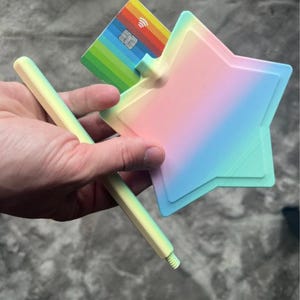 Może przedstawiać: Pastelowy, tęczowy ściągacz do kart w kształcie gwiazdy z pasującym pastelowo-ż&oacute;łtym uchwytem w kształcie patyczka. Gwiazda posiada szczelinę do włożenia karty płatniczej, w kt&oacute;rej obecnie znajduje się wielokolorowa karta w paski.