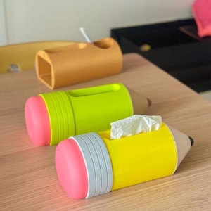 May include: Three pencil-shaped desk organisers and tissue holders. One is tan, one is lime green with a pink eraser end, and one is yellow with a grey band and pink eraser end. The yellow holder contains a white tissue protruding from the top.