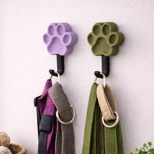 May include: Two wall-mounted hooks featuring paw-shaped tops in light purple and olive green. Each hook has a black metal arm and a curved base. A purple and gray harness hangs from the purple hook, while a green and beige leash hangs from the green hook.