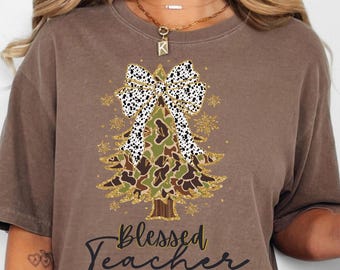 May include: A brown short-sleeve t-shirt featuring a graphic of a Christmas tree with a camouflage pattern, a white and black leopard print bow, and gold glitter accents. The text "Blessed Teacher" is printed in black cursive script below the tree.