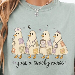 Könnte beinhalten: Ein salbeigrünes T-Shirt mit einer Grafik von vier geisterhaften Enten als Krankenschwestern, die bunte Patchwork-Muster tragen. Der Text darunter lautet "just a spooky nurse" in schwarzer Schreibschrift, ergänzt durch kleine Sterne, einen Mond und Herzen.