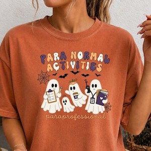 Peut inclure: Un t-shirt à col rond orange brûlé avec un motif de trois fantômes et le texte "PARA NORMAL ACTIVITIES" et "paraprofessional". Les fantômes sont représentés tenant un presse-papier, une tasse de café, un talkie-walkie et des livres.