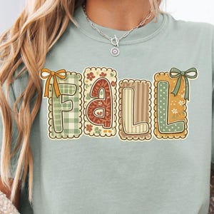 Può includere: Una t-shirt a maniche corte color verde salvia con la parola "fall" in grandi lettere in stile blocco. Ogni lettera è incorniciata da un bordo smerlato e decorata con diversi motivi, tra cui vichy verde, fiori, righe e pois, con fiocchi.