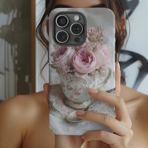Op de afbeelding: Een telefoonhoesje met een bloemenontwerp. De hoes toont een theekopje gevuld met roze rozen en andere bloemen. Het theekopje staat op een kanten placemat met een lepel. De hoes heeft een grijze achtergrond.