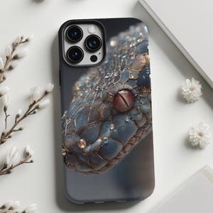 Op de afbeelding: Een smartphonehoesje met een gedetailleerde afbeelding van een slangenkop. Het ontwerp toont blauwe en bruine schubben met waterdruppels en een rood en bruin oog. De hoes heeft een zwarte cameramodule. De achtergrond is wit.