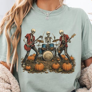 May include: A sage green t-shirt featuring a graphic of three skeletons playing musical instruments, including a red electric guitar, a drum set, and a black bass guitar, surrounded by orange pumpkins on the ground in a field.