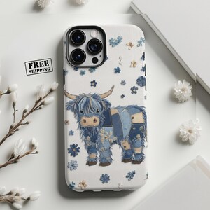 May include: A white phone case featuring a Highland cow illustration with blue denim patchwork and floral accents. The case has a black camera module and is decorated with small blue and beige flowers. The words "FREE SHIPPING" are printed on the case.