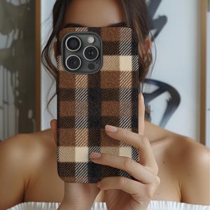 May include: A brown, black, and cream plaid phone case. The case has a textured appearance and covers the back of a smartphone. The camera lenses are visible. The case is held in front of a person.