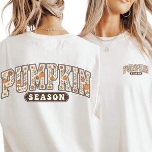 May include: A white t-shirt featuring the text PUMPKIN SEASON in a large arched font on the back, filled with orange pumpkins, brown leaves, and small flowers. A smaller version of the same text and floral pumpkin graphic is printed on the front left chest.