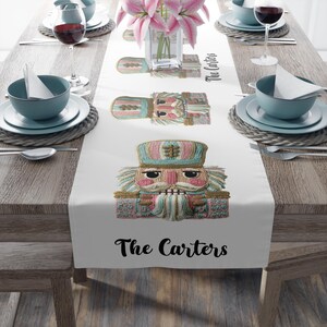 May include: White table runner featuring three embroidered nutcracker figures in pastel colors. The text "The Carters" is printed in black at the bottom. The runner is set on a wooden table with place settings and flowers.