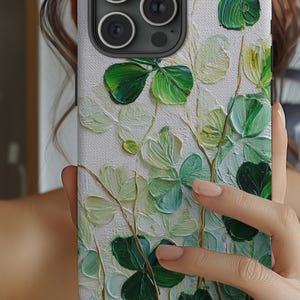 May include: A white phone case with a textured, painted design of green and light green shamrocks. The shamrocks have a 3D effect, with visible brushstrokes. The case has a black camera module.