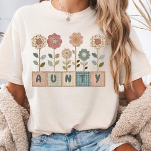 Könnte beinhalten: Cremefarbenes T-Shirt mit floralem Design und dem Wort "AUNTY" in Patchwork-Buchstaben. Die Blumen sind in verschiedenen Farben und Mustern gehalten, mit grünen Stielen und Blättern. Ein lässiges und bequemes Oberteil.