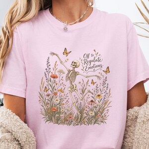 Puede incluir: Una camiseta rosa claro con un estampado gráfico de un esqueleto bailando en medio de un campo de flores silvestres y mariposas. El texto "Off to Regulate Emotions" aparece en una fuente cursiva oscura sobre el esqueleto, junto a un pequeño corazón.
