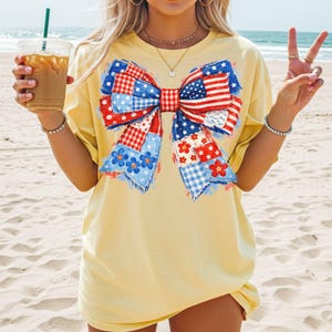 May include: A pale yellow t-shirt featuring a large, central graphic of a bow made from a patchwork of red, white, and blue patterns, including stars, stripes, gingham, and floral prints, with frayed edges on the bow's ribbon tails.