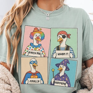 May include: A sage green t-shirt featuring a four-panel graphic print of cartoon ducks dressed as a clown, a pirate, a superhero, and a wizard. Each duck holds a sign with the text "PINCH ME", "WHEN IT", "FINALLY", and "HAPPENS" respectively.