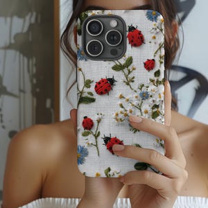 Pode incluir: Uma capa de telefone branca com bordados de joaninhas vermelhas, margaridas brancas e amarelas, flores azuis e folhas verdes. A capa tem um recorte preto para a lente da câmera.