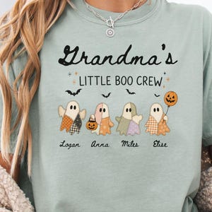 Puede incluir: Una camiseta verde salvia con el texto Grandma's LITTLE BOO CREW en negro. Debajo del texto hay cuatro ilustraciones de fantasmas con patrones de patchwork en naranja, beige y morado, sosteniendo un cubo de calabaza y un globo, con los nombres Logan, Anna, Miles, Elise.