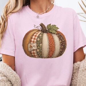 May include: A light pink t-shirt featuring a large, central graphic of a pumpkin constructed from various fabric patches. The pumpkin design includes segments of brown, orange, and green patterns, including gingham, floral, and herringbone textures.